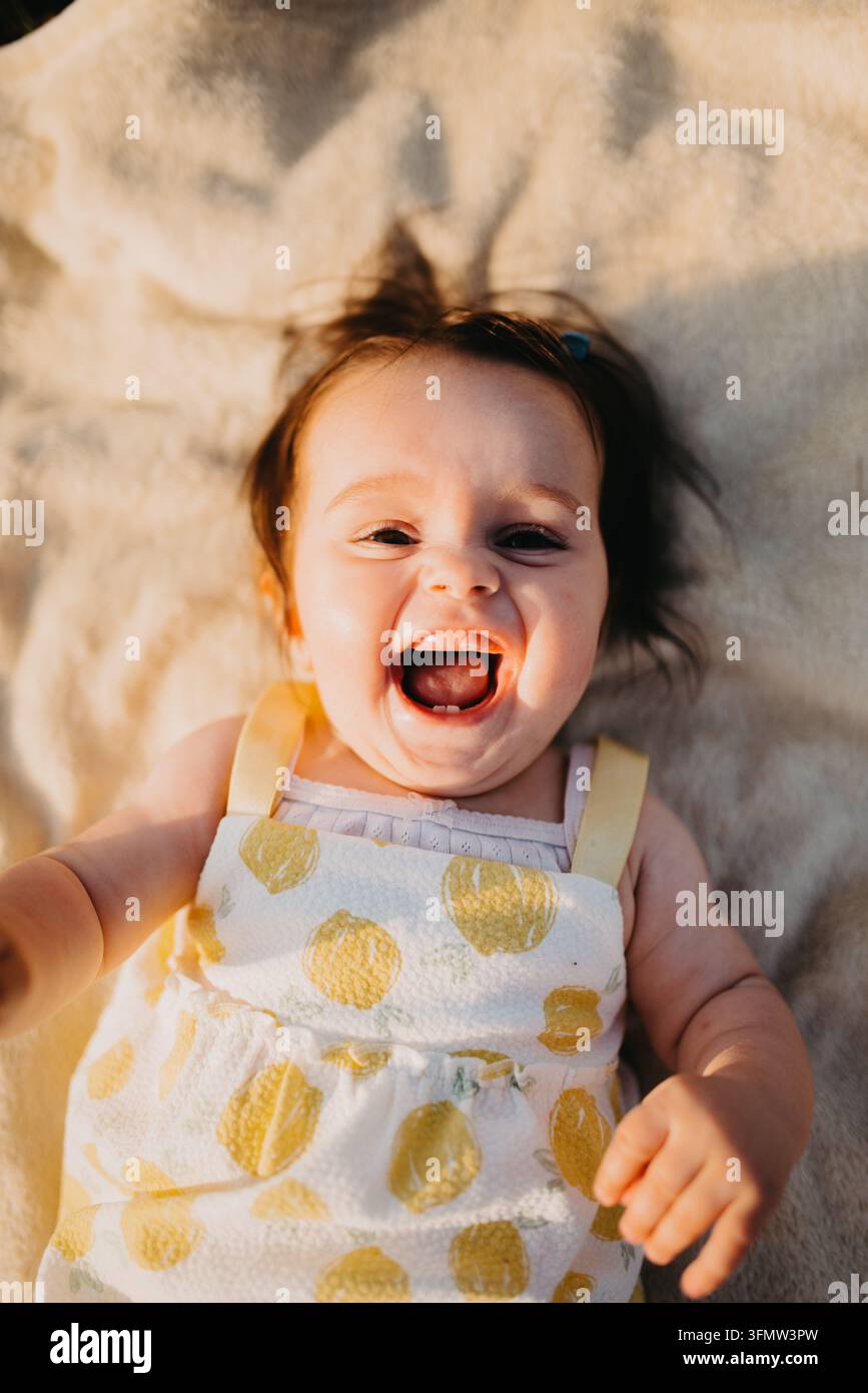 Bambino che ride sdraiato su una coperta con indosso un vestito giallo a stampa limone, ritratto gioioso di un neonato Foto Stock