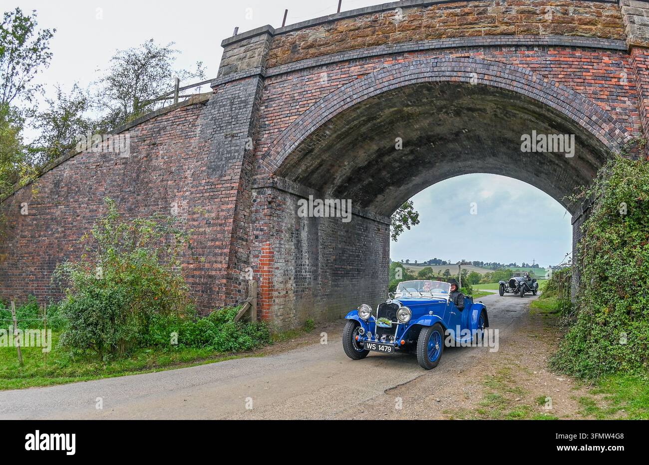 Una classica auto vintage a cielo aperto Triumph FS percorre una strada sterrata rurale passando sotto un ponte storico in mattoni ad arco. Foto Stock