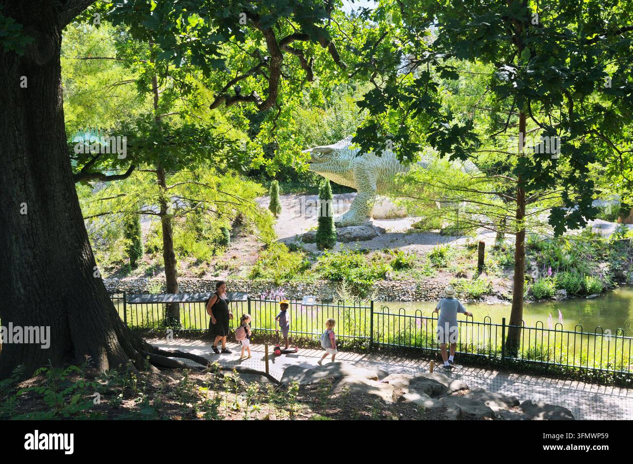 Adulti e bambini che esplorano le sculture di modelli di dinosauri vittoriani nel Crystal Palace Park. Megalosaurus incombente sullo sfondo. Londra UK Dinosauri Foto Stock