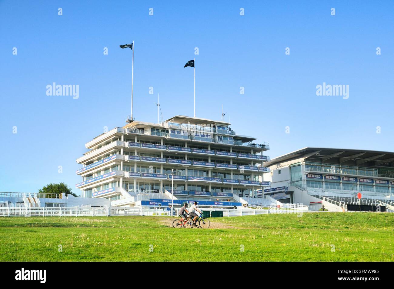 Due uomini in bicicletta davanti all'Epsom Grandstand Enclosure, Stand Queen Elizabeth II, Ippodromo di Epsom Downs (Casa del Derby), Surrey, Inghilterra, Regno Unito.  Ciclisti Foto Stock