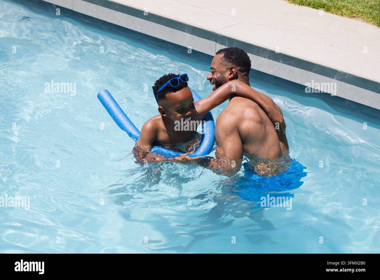 Padre afroamericano che tiene suo figlio adolescente che gioca a biliardo in cortile, indossando pantaloncini, occhiali protettivi e una canna di piscina Foto Stock