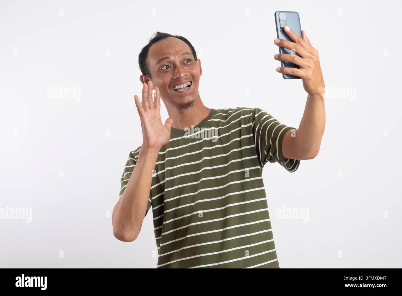 Giovane uomo positivo con maglietta verde che saluta con la mano durante una videochiamata tramite telefono cellulare, isolato su uno sfondo bianco da studio Foto Stock