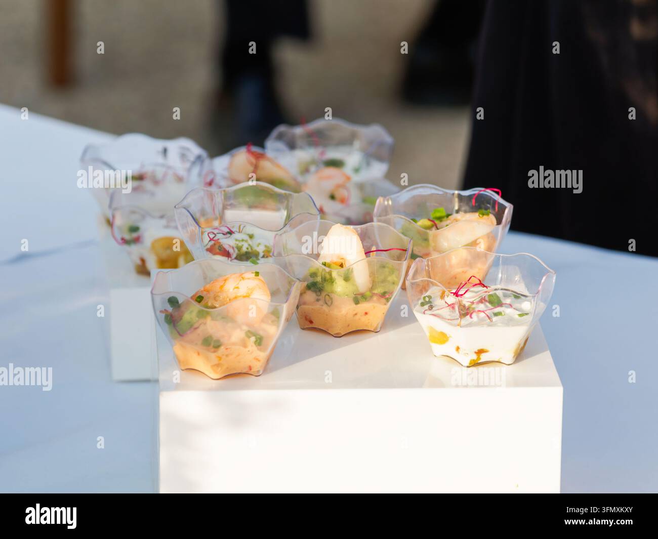 Vari antipasti serviti in piccole coppette di plastica trasparente su una superficie bianca durante un evento all'aperto. Foto Stock