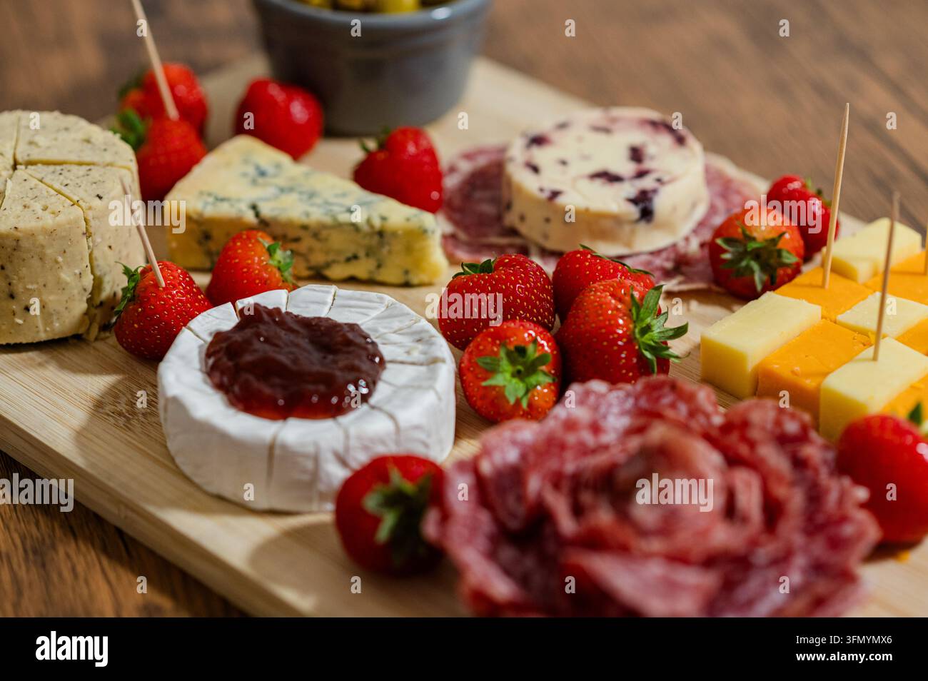 Tagliere di legno disposto con formaggi assortiti, salumi stagionati, fragole fresche, marmellata e stuzzicadenti per uno spuntino appetitoso, un antipasto o una gougère Foto Stock