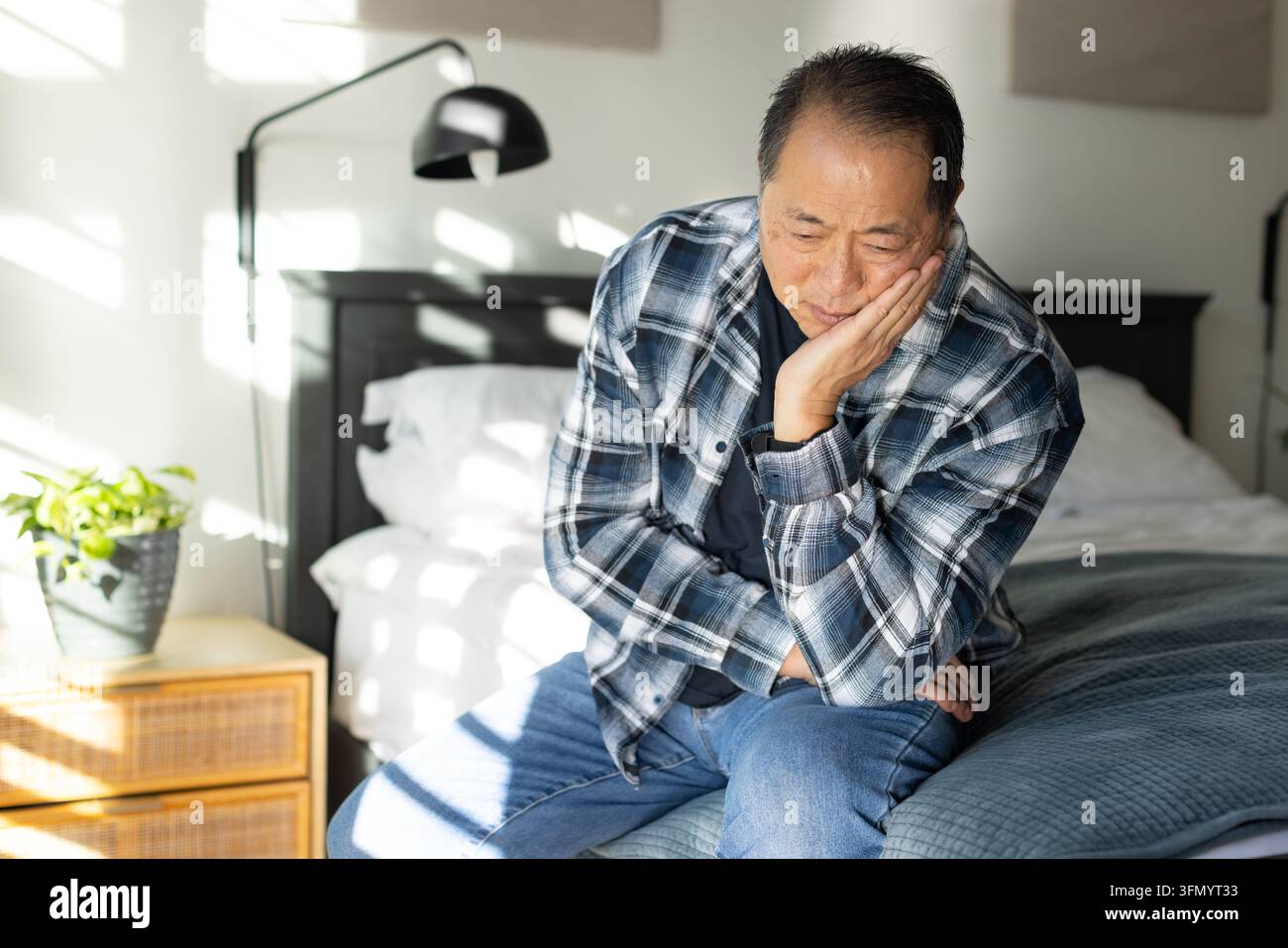 Uomo asiatico anziano seduto sul letto, indossa una camicia a quadri, appoggia la guancia sulla mano, la luce del sole che entra Foto Stock