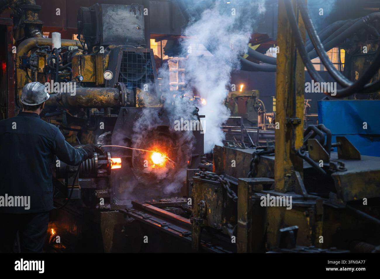 Un operaio siderurgico lavora accanto alla lavorazione di tubi d'acciaio caldi sulla linea di produzione di una fabbrica di tubi industriali. Foto Stock