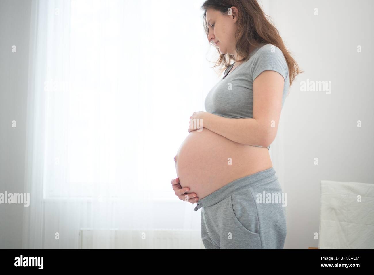 Donna incinta con le mani sullo stomaco in profilo laterale rilassato che abbraccia il bagliore naturale della gravidanza in uno studio caldo. Madre in attesa con espressione serena e postura comoda in un interno bianco minimale. Foto Stock