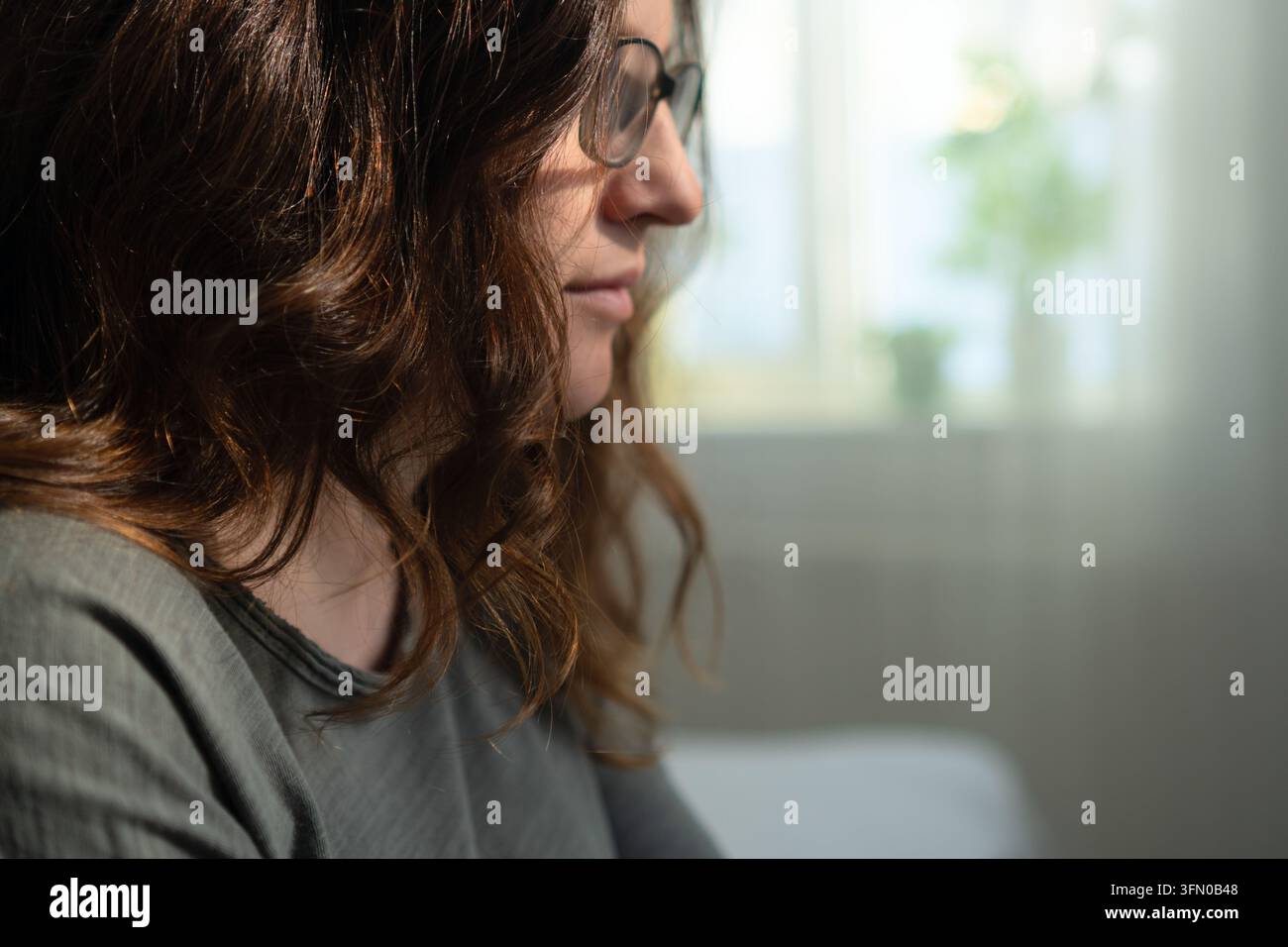 Bruna misteriosa nasconde il suo volto dietro ciocche scure e occhiali in un ritratto in primo piano al chiuso. La donna riservata si allontana, il suo sguardo enigmatico nascosto nella luce soffusa su uno sfondo sfocato. Foto Stock