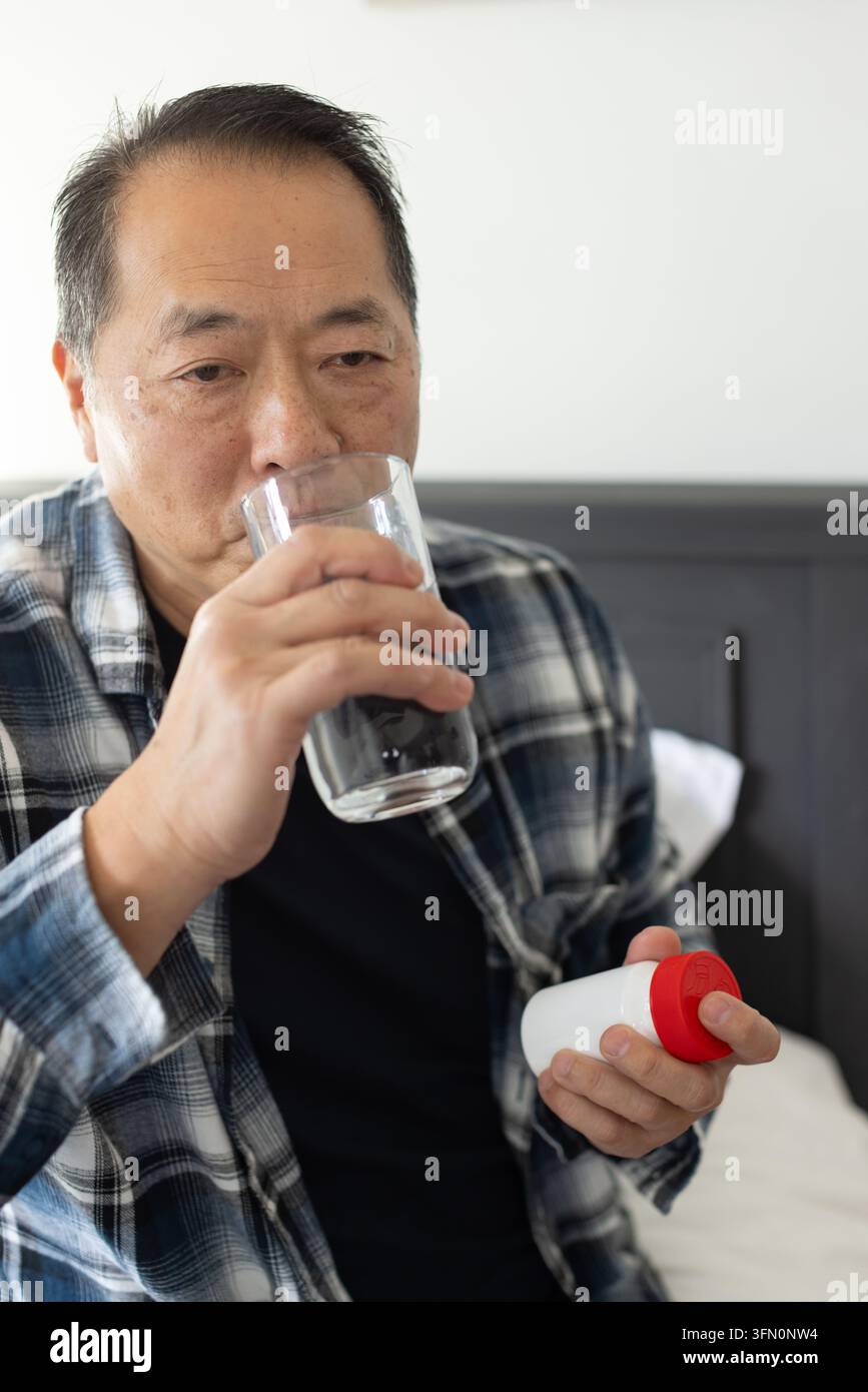 Uomo asiatico anziano in camicia a quadri seduto sul letto, che tiene una bottiglia con tappo rosso e beve da un bicchiere trasparente Foto Stock