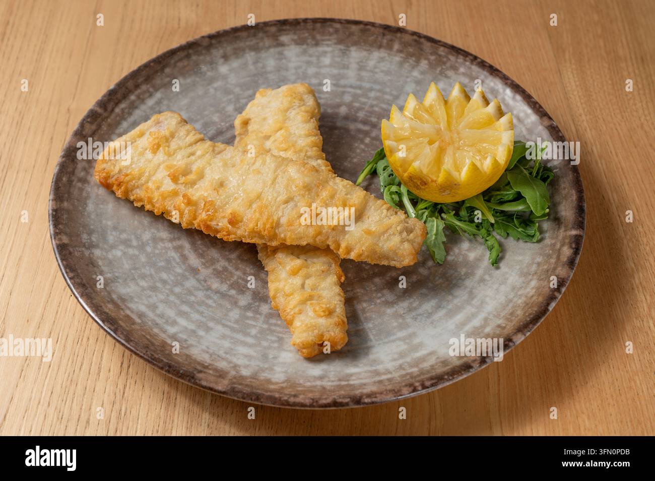 Filetti di pesce impanati e fritti serviti con limone e rucola su un piatto di ceramica rustico Foto Stock