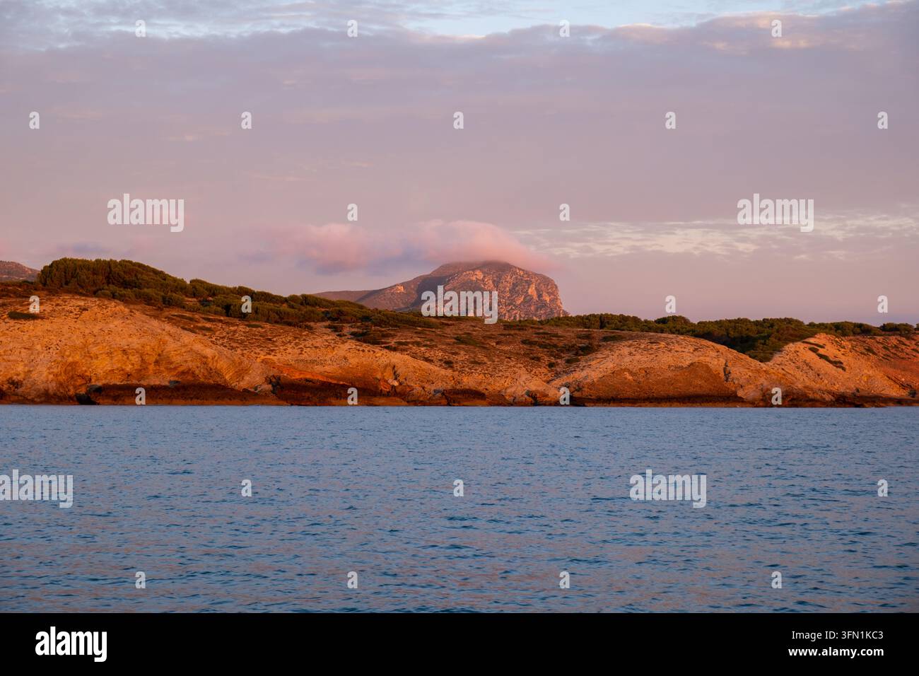 La luce calda dell'ora dorata avvolge scogliere rosse frastagliate e un picco montuoso drammatico sulla costa di Maiorca, ideale per concetti di viaggio e natura. Foto Stock