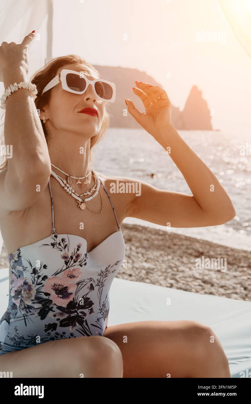Donna spiaggia tramonto persona elegante in costume da bagno floreale e occhiali da sole bianchi seduta su un lettino da spiaggia durante le vacanze estive Foto Stock