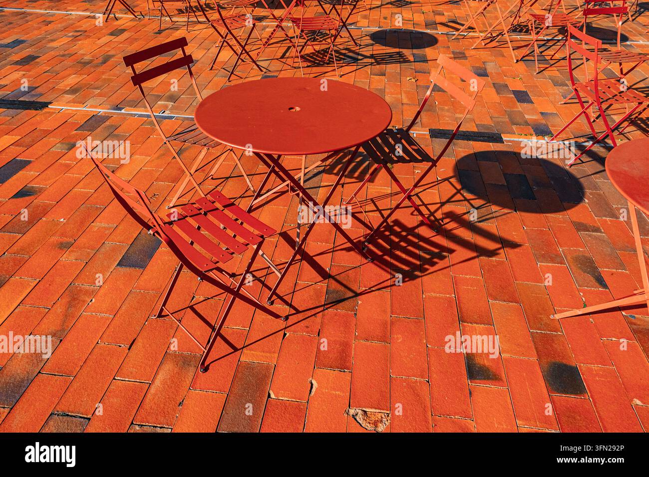 Sedie e tavoli da caffè in metallo rosso proiettano ombre geometriche audaci su pavimenti in mattoni arancioni sotto la luce del sole, senza persone presenti Foto Stock