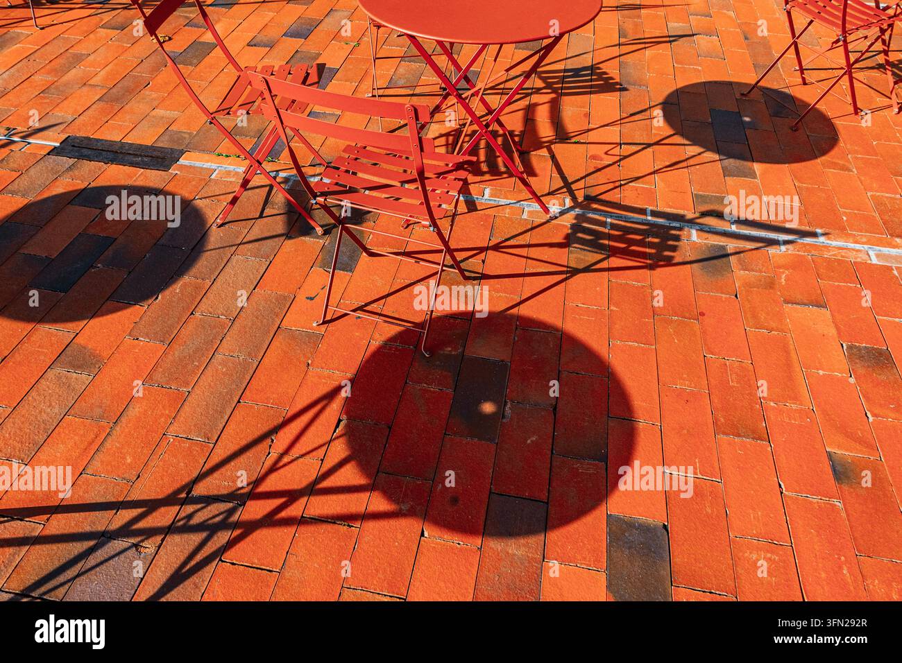 Sedie e tavoli da caffè in metallo rosso proiettano ombre geometriche audaci su pavimenti in mattoni arancioni sotto la luce del sole, senza persone presenti Foto Stock