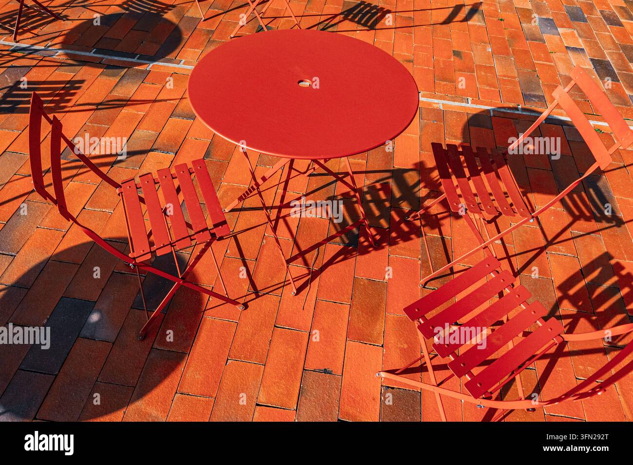 Sedie e tavoli da caffè in metallo rosso proiettano ombre geometriche audaci su pavimenti in mattoni arancioni sotto la luce del sole, senza persone presenti Foto Stock