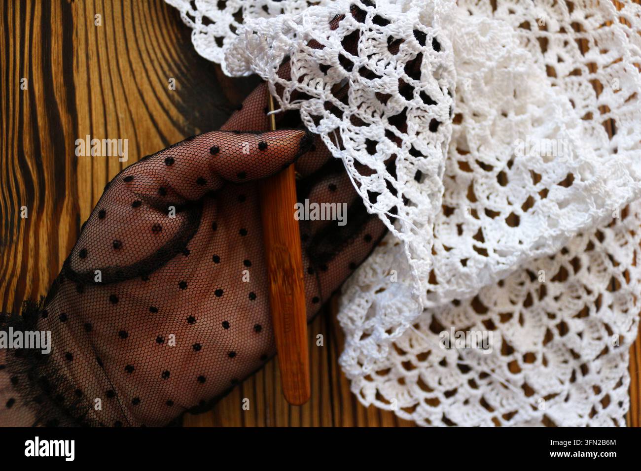 Mano che indossa un guanto a rete a pois neri e tiene il bordo ondulato di uno scialle di pizzo all'uncinetto bianco su uno sfondo di legno scuro. Eleganza, vintage fa Foto Stock