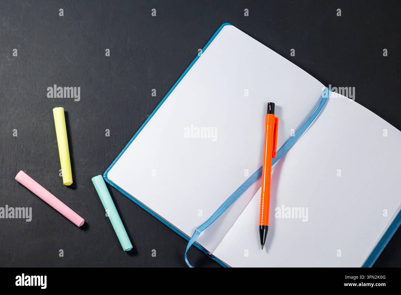 Un quaderno blu aperto e vuoto con una matita arancione brillante all'interno, posizionato accanto a pezzi di gesso colorati su una superficie di legno scuro, perfetto per concetti di ritorno a scuola. Foto di alta qualità Foto Stock