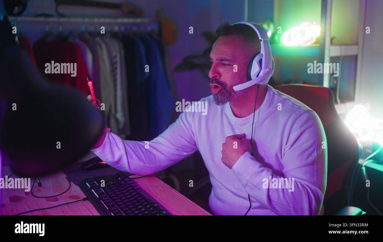 Uomo con auricolari tiene uno smartphone e gesticola con la mano in un setup da gioco in studio; gioia del gioco online. Foto Stock