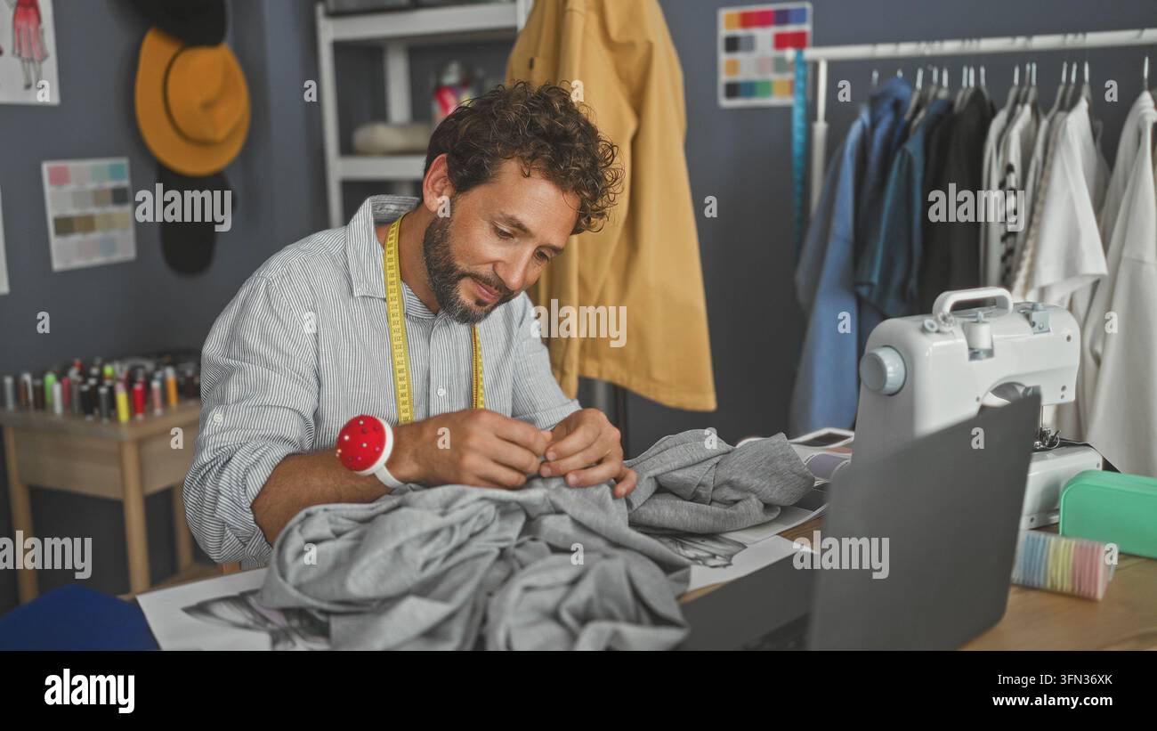 Uomo che cuce a mano un indumento grigio su un tavolo da studio con macchina da cucire, metro e laptop visibili; attenzione alla lavorazione artigianale. Foto Stock