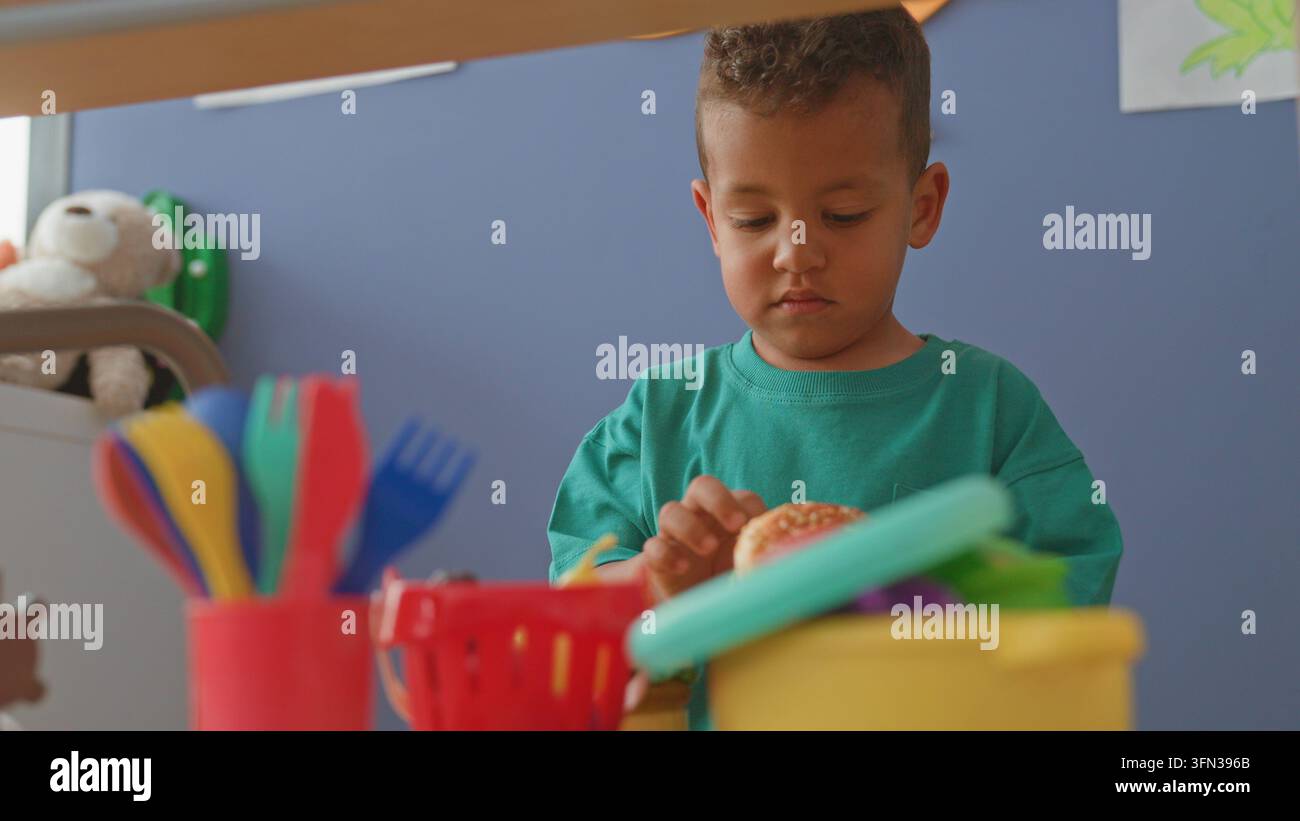 Ragazzo che tiene un hamburger giocattolo e gioca con utensili di plastica a un tavolo da gioco; concentrazione silenziosa. Foto Stock