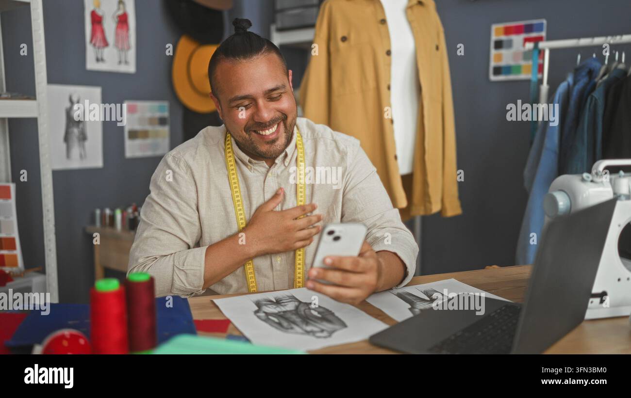 Sarto maschile sorridente, tiene uno smartphone con le mani e un metro a nastro intorno al collo in uno studio di design; gioia creativa. Foto Stock