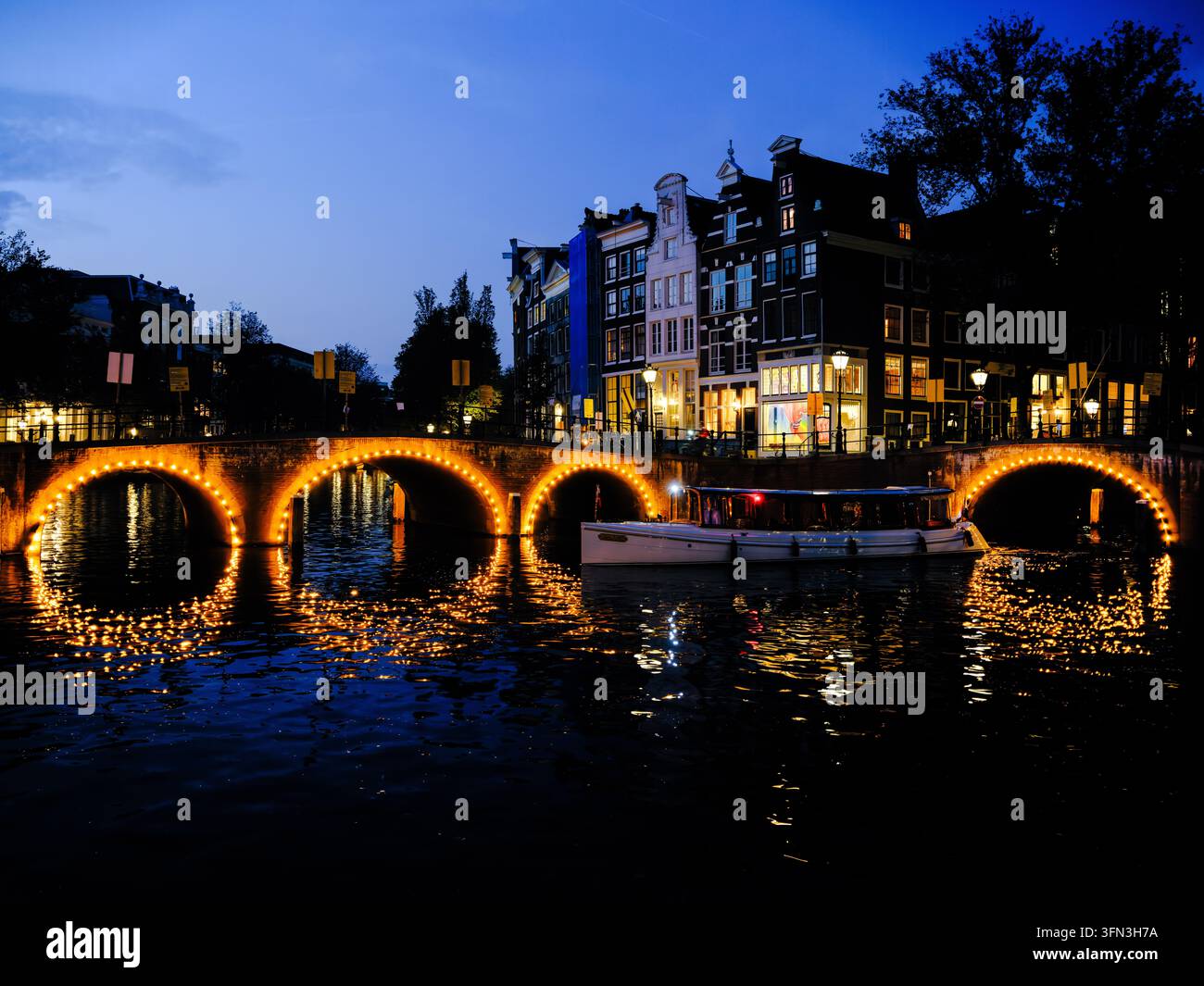 Un ponte ad arco illuminato e una barca turistica si riflettono nell'acqua all'ora blu ad Amsterdam, Paesi Bassi Foto Stock