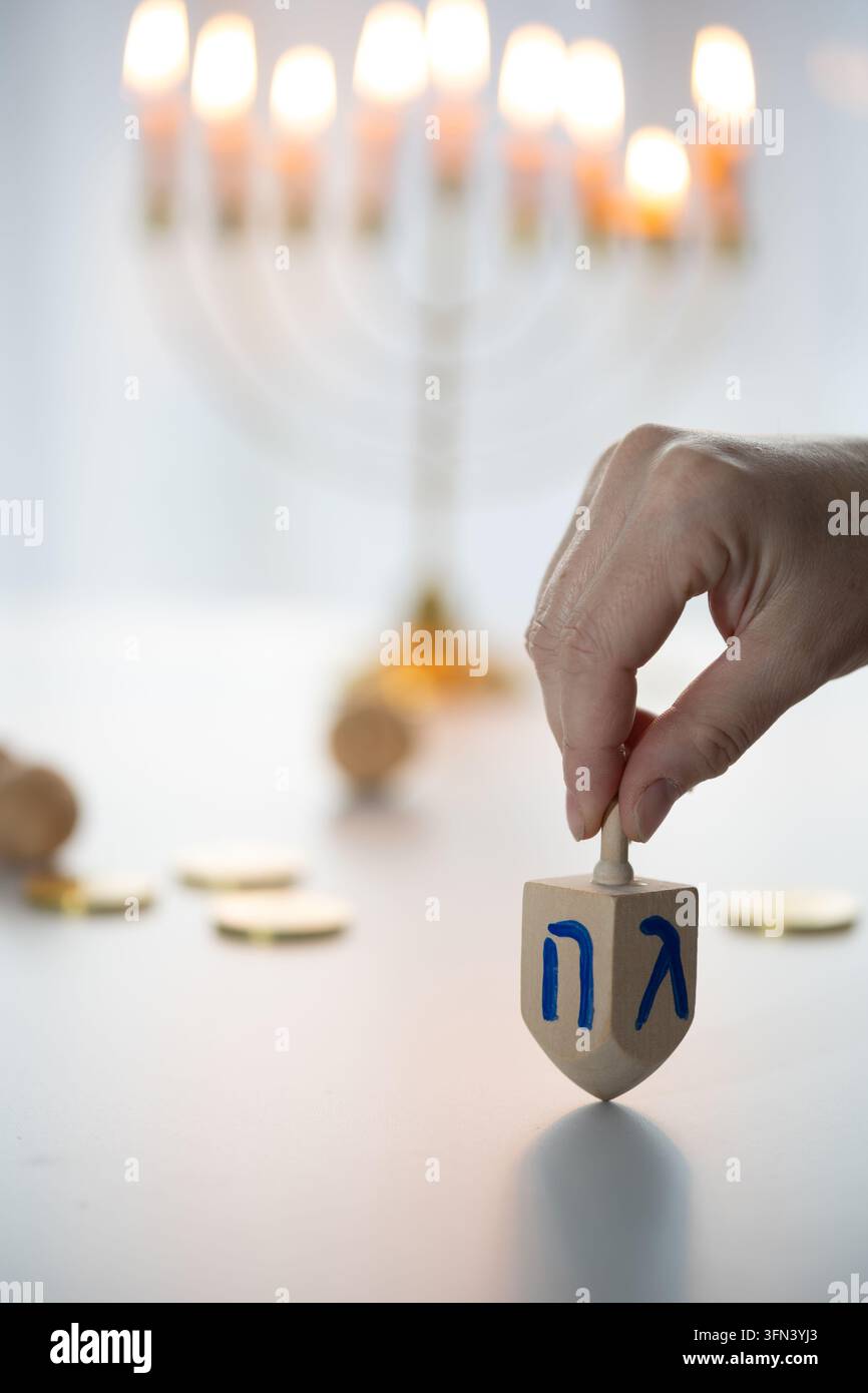 Primo piano di una mano che tiene un dreidel di legno su un tavolo bianco, mostrando lettere ebraiche mentre il giocoliere poggia in posizione verticale, con una luce naturale soffusa Foto Stock