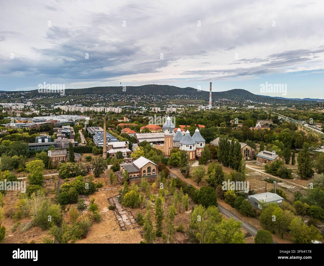 Vista aerea della storica fabbrica di gas di Obuda, con edifici industriali in mattoni, torri e camini tra gli alberi, sviluppo urbano e le colline di Buda a B Foto Stock