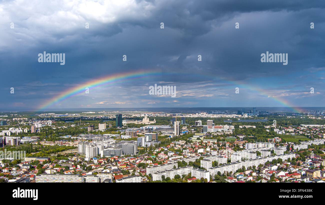 Vista aerea di un vivace arcobaleno che si inarca sulla città di Bucarest, con blocchi residenziali e spazi verdi sotto un drammatico cielo tempestoso in Foto Stock
