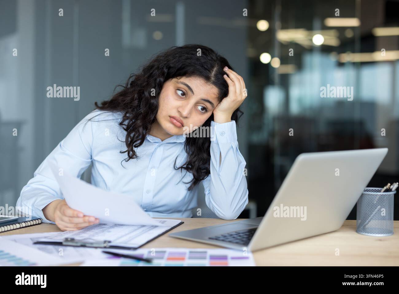 Giovane donna d'affari con capelli ricci scuri seduta alla scrivania dell'ufficio, tiene documenti e sembra stanca e sopraffatta mentre vive stress lavorativo e burnout Foto Stock