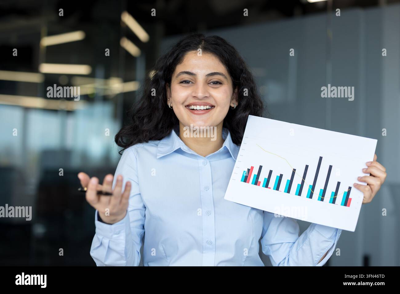 Giovane donna d'affari indiana che presenta grafici a barre finanziari e dati con un sorriso sicuro, condividendo approfondimenti sulla crescita e lo sviluppo del business in un ambiente d'ufficio moderno Foto Stock
