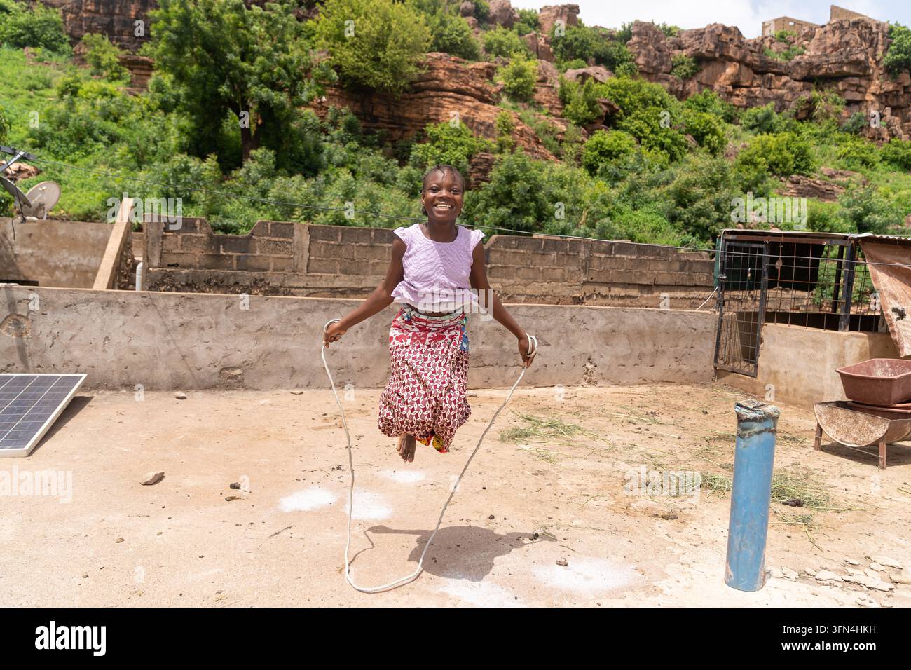 Ragazza africana sorridente salta la corda all'aperto vicino a un pannello solare, combinando gioco infantile, esercizio fisico, empowerment delle ragazze, energia rinnovabile e azione climatica Foto Stock