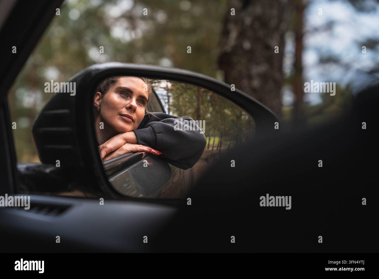 Viaggiatrice pensierosa che appoggia la testa sulla portiera dell'auto riflessa nello specchietto retrovisore in mezzo a un paesaggio boschivo Foto Stock