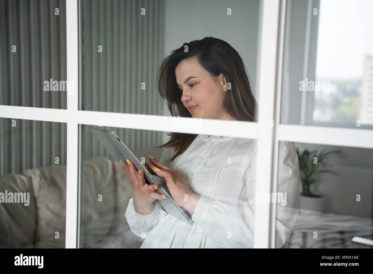 Donna che legge contenuti digitali su un tablet accanto a una finestra luminosa di casa. Apprendimento online autonomo, stile di vita digitale ed educazione a distanza. Foto Stock