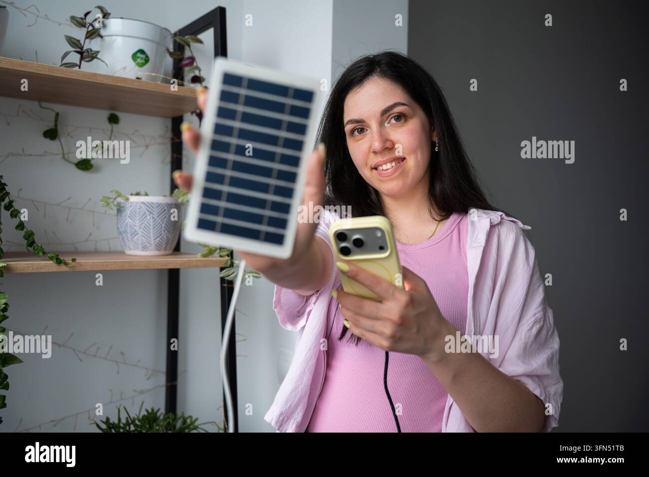 Giovane donna che tiene un pannello solare portatile e uno smartphone in un interno domestico. Ricarica solare, vita sostenibile e tecnologia di energia rinnovabile. Foto Stock