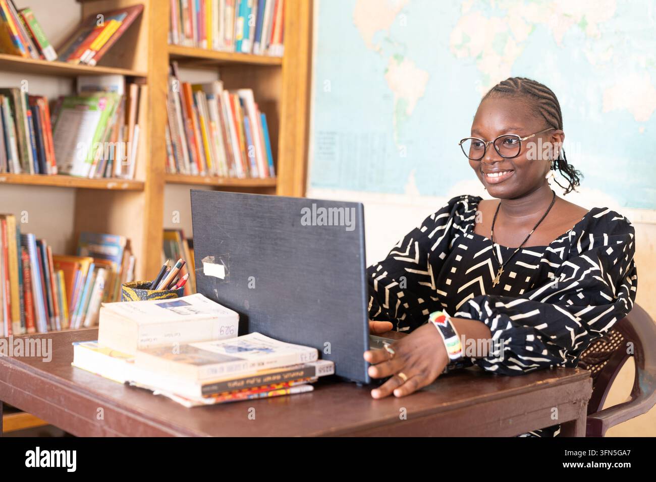 Donna africana sorridente utilizza uno smartphone in una biblioteca, circondata da libri e materiale di studio, simboleggiando l'alfabetizzazione digitale, l'istruzione e l'emancipazione delle donne Foto Stock