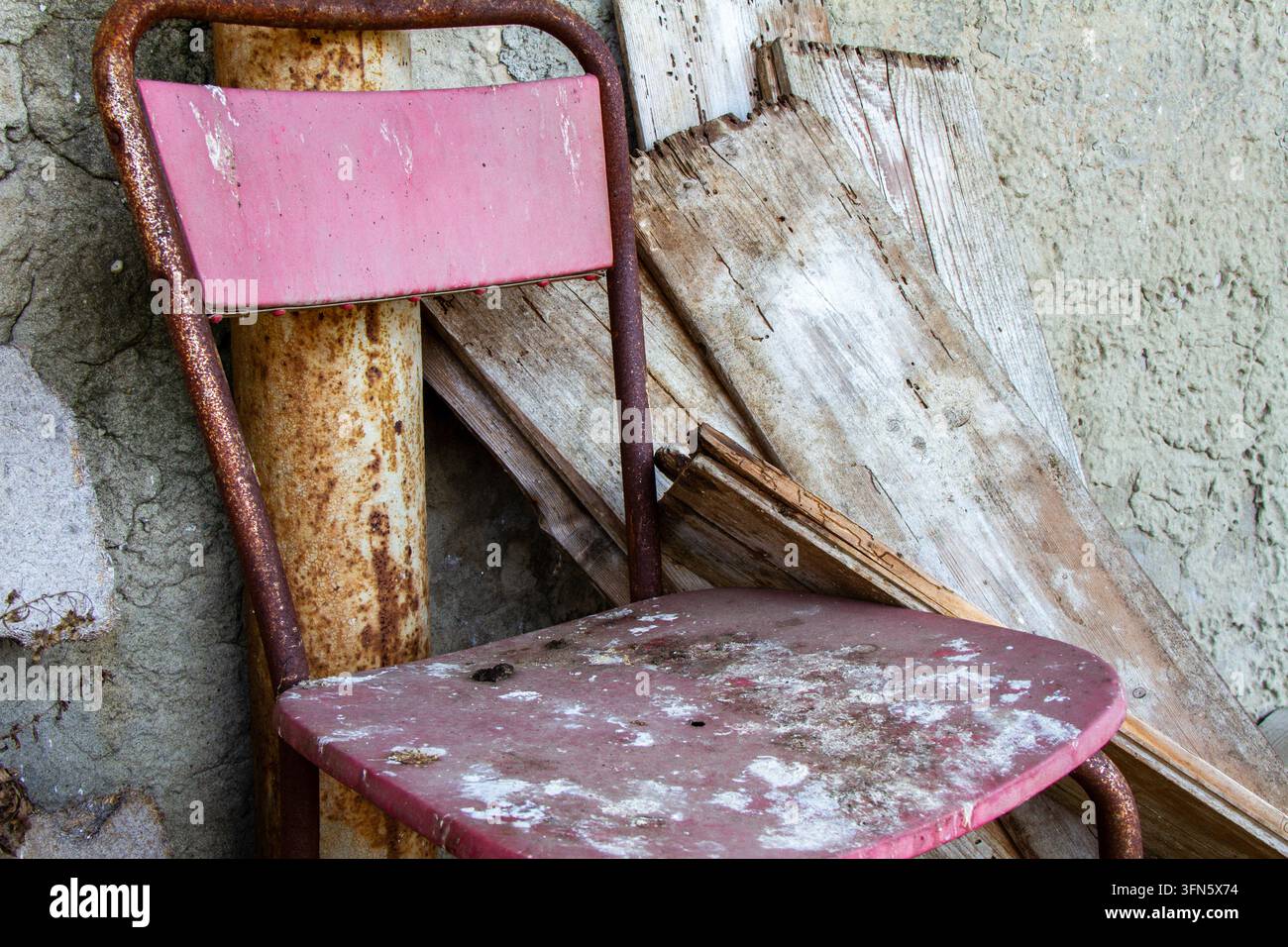 Un primo piano mostra una sedia in metallo rosa sporca e arrugginita appoggiata a un muro di cemento ruvido. Accanto ci sono un tubo metallico corrose e un vecchio w eroso Foto Stock