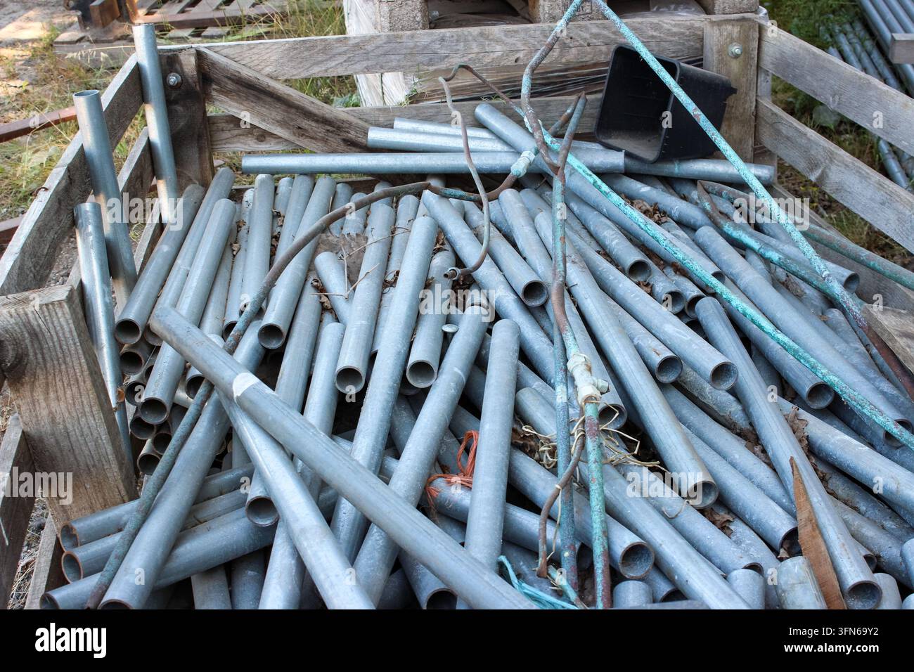 Pila di tubi metallici galvanizzati e ossidati di varie dimensioni impilati in una cassa di legno rustica all'aperto, accanto a un vaso di plastica nero. Foto Stock