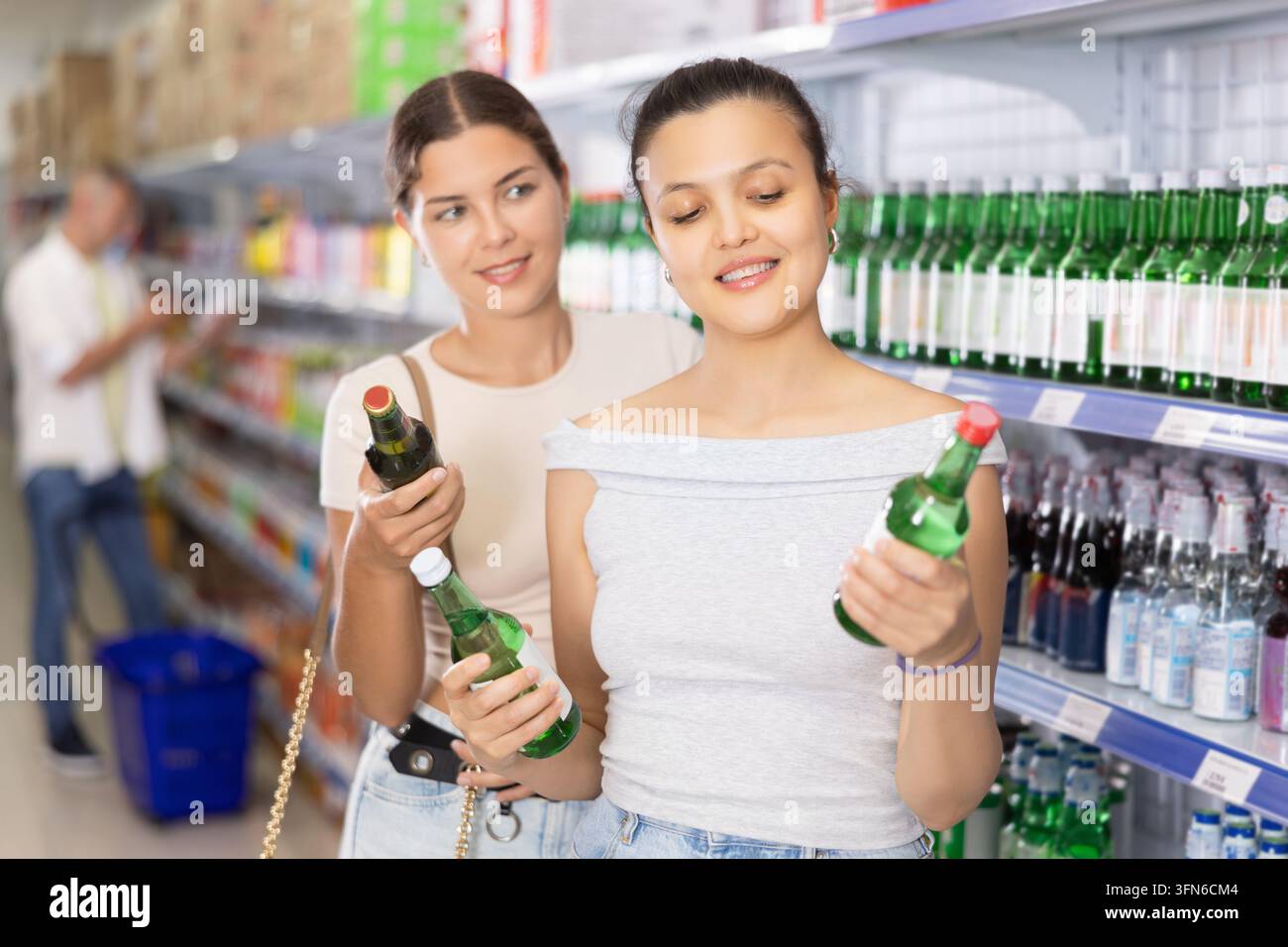 Due giovani donne acquirenti in piedi accanto agli scaffali con varie bevande alcoliche nel supermercato, esaminano le etichette sulle bottiglie e discutono amichevolmente sulla scelta Foto Stock