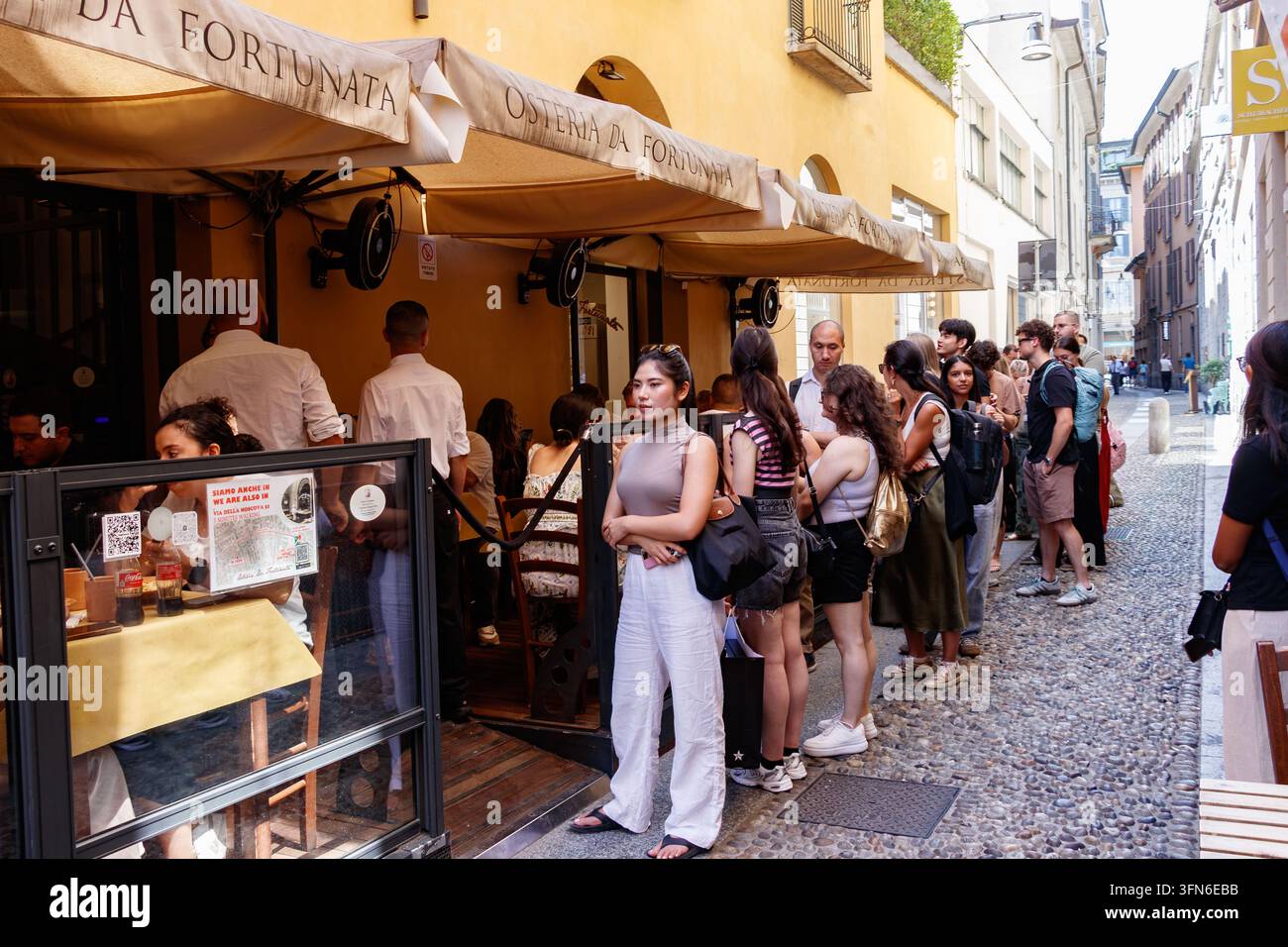 I clienti fanno la fila fuori dal popolare ristorante Osteria da Fortunata con una donna che indossa pantaloni bianchi in una strada di Milano Foto Stock