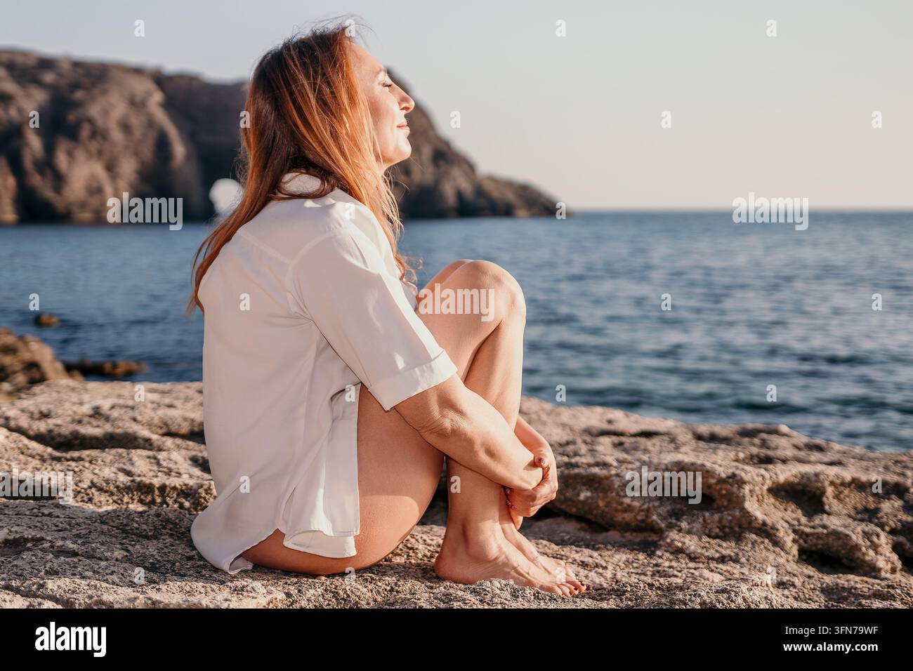 Donna che si rilassa seduta su una riva rocciosa vicino al mare all'alba per godersi una meditazione tranquilla e momenti di quiete Foto Stock