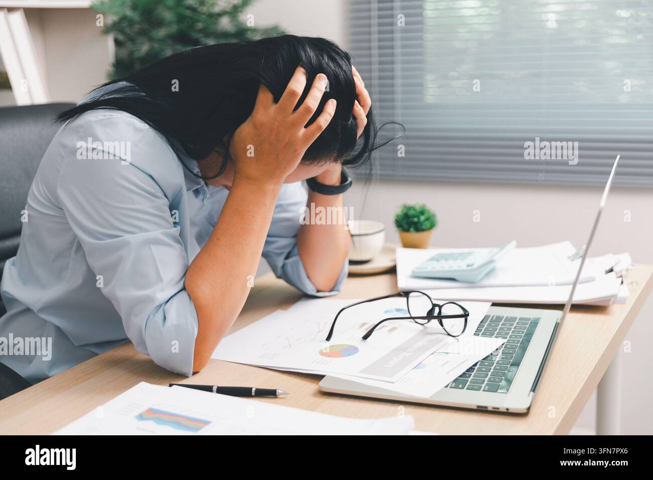 Donna d'affari sopraffatta che tiene la testa a una scrivania con laptop, calcolatrice e rapporti, mostrando burnout, stress lavorativo, pressione delle scadenze, w finanziaria Foto Stock