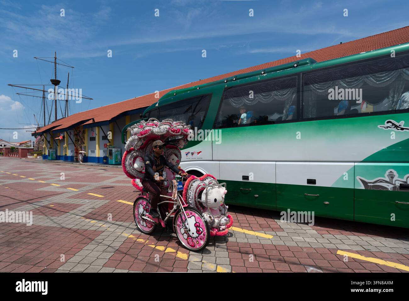 Melaka, Malesia - Settembre 2026: Un guidatore di trishaw locale pedala un rickshaw a tre ruote a tema Hello Kitty decorato in modo vivace davanti a un autobus turistico Foto Stock