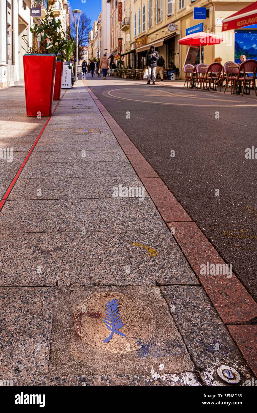 Primo piano del simbolo metallico a forma di foglia di palma nella vecchia città di Cannes con turisti sfocati e negozi sotto un cielo blu Foto Stock