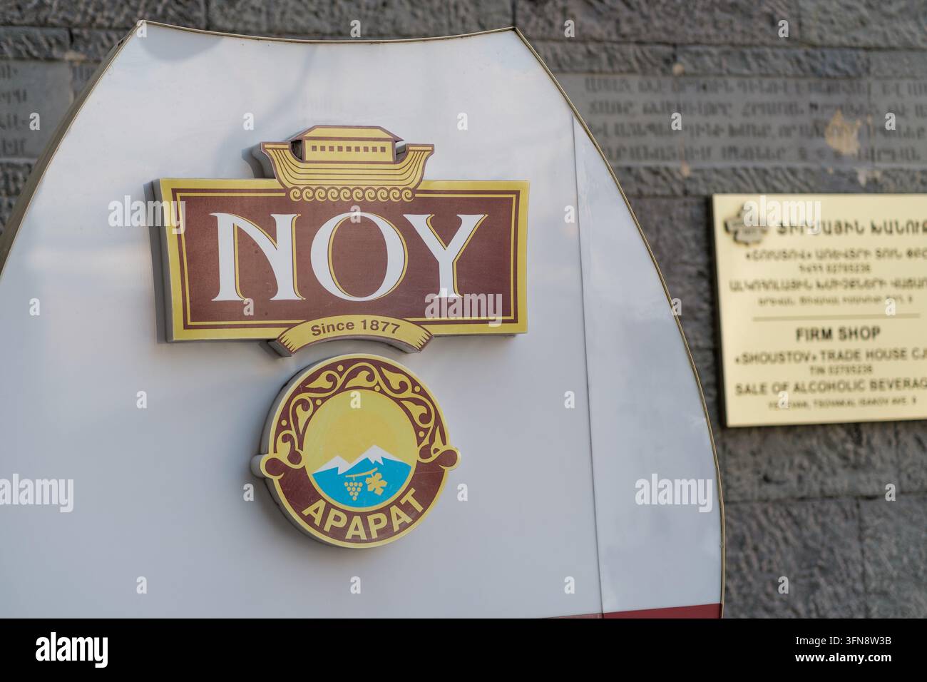Yerevan, Armenia - 27 agosto 2026: Il logo Noy brandy e l'emblema di Ararat appaiono su un cartello a forma di scudo accanto a una targa informativa al Noah brandy Foto Stock
