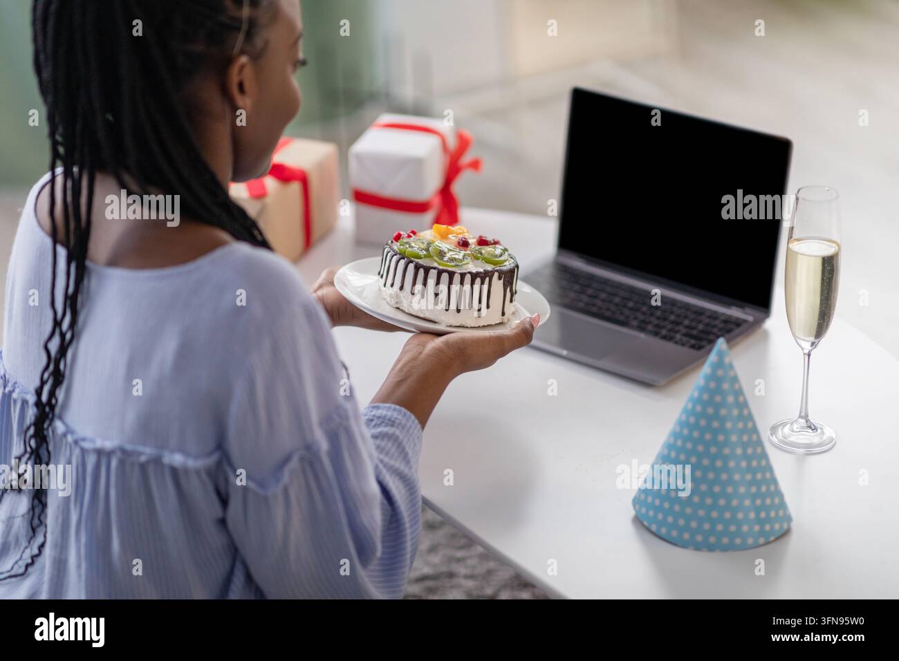Donna afroamericana che tiene una torta di compleanno davanti a un laptop con schermo vuoto, ripresa sopra la spalla. Donna nera irriconoscibile che mostra una bella C Foto Stock