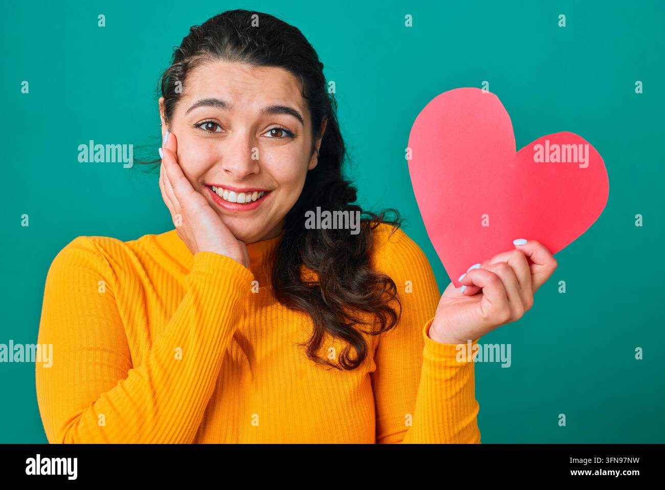 Donna sorridente che tiene un cuore di carta rosso mostrando amore e sorpresa su uno sfondo turchese Foto Stock