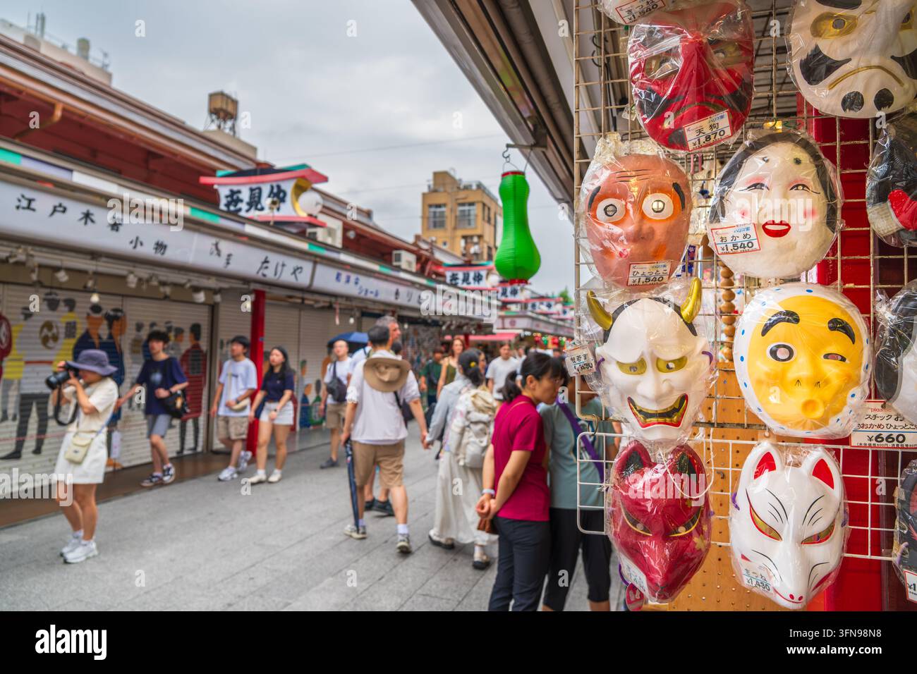 Tokyo, Giappone – 7 agosto 2025: Maschere tradizionali giapponesi colorate sono esposte su una rastrelliera in un mercato di strada ad Asakusa, Tokyo con visitatori nel Foto Stock