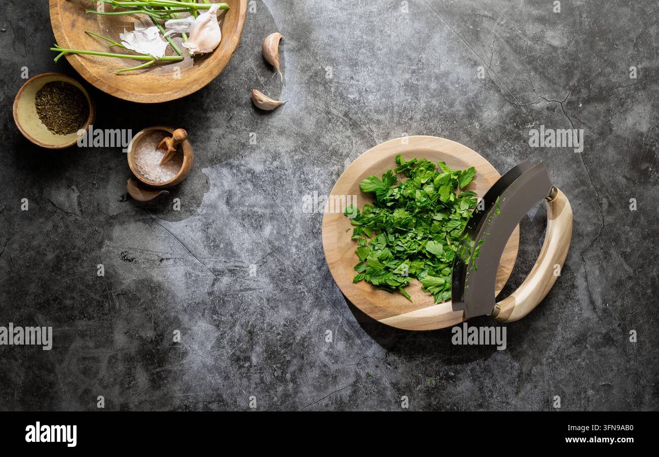 Erbe verdi tritate in una piccola ciotola di legno con un coltello mezzaluna e altri ingredienti da cucina su una superficie rustica Foto Stock