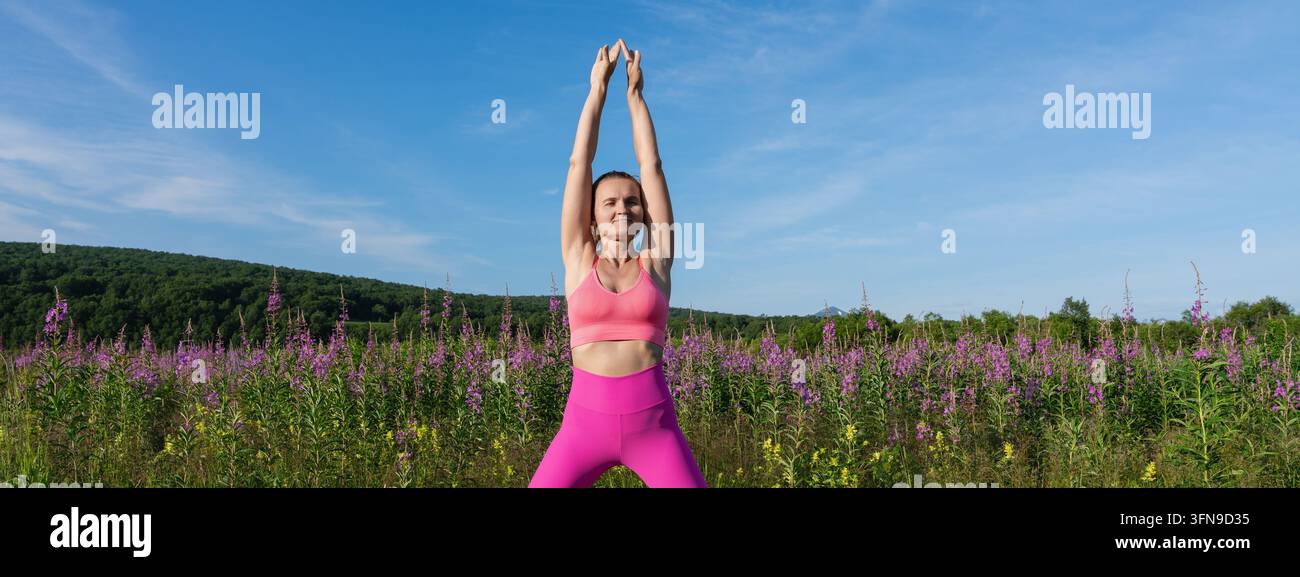 Donna ben formata in abbigliamento sportivo rosa che allunga le braccia sopra la testa in un prato fiorito con colline e cielo azzurro. Allenamento di yoga e pilates all'aperto. Fitness, salute Foto Stock